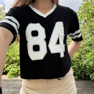 Sportig t-shirt topp - Svart, stretchig och croppad t-shirt/topp med tryck ”84” samt ränder på armar i silver och vit från Forever 21. Storlek: M. Material: 94% viskos + 6% elastan. :)