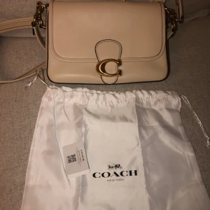 Beige handväska från Coach - En elegant beige handväska från Coach med en gyllene spänne i form av ett 'C'. Väskan har en justerbar axelrem och kommer med en vit förvaringspåse med Coach-logotypen.