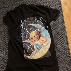 En svart t-shirt från Gildan med ett tryck av en ängel som sitter på en måne, omgiven av blixtar. T-shirten har korta ärmar och är i mjukt material.