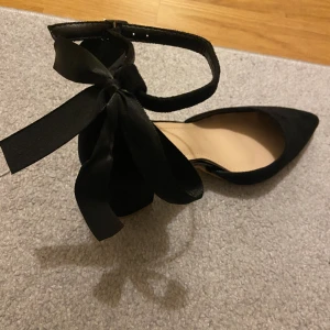 Klackskor, svarta med roset  - Säljer ett par helt oanvända klackskor i svart färg och med en roset back vid hälen. Säljer pga köpt fel storlek, dvs storlek 37. Perfekta till kommande bal eller annan festlighet. Pris: 130kr