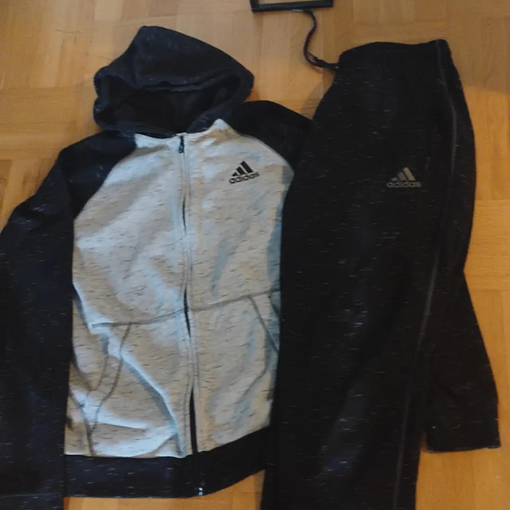 Svart och grå Adidas munkaka med huva och dragkedja samt fickor fram.  Tillhörande byxor svarta Adidas stl L, ( 500 kr för båda el 300 kr st Adidas jakobsskolan. Hoodies.