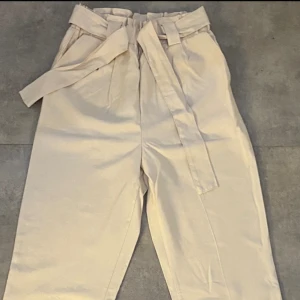 Kostymbyxor- beige - Klassiska kostymbyxor i färgen beige med  knytband och resår vid midjan. Byxan har raka ben och är använda endast en gång. Ny utan prislapp.