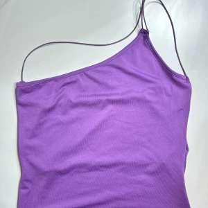 Topp med djup rygg - Den här toppen har ett one-shoulder-look med 2 tunna, ojusterbara, elastiska axelband. Den har en draperad djup rygg med en tunn remdetalj och en croppad längd. Den här toppen har ett mjukt, stretchigt material. Den här toppen kommer i lila.