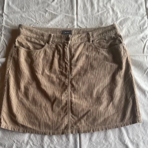 Beige manchesterkjol från MEXX - En beige manchesterkjol från MEXX i storlek L. Kjolen har en klassisk design med fram- och bakfickor samt en knapp och dragkedja framtill. Materialet är ribbat och ger en texturerad känsla.