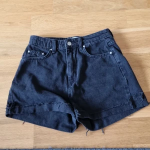 Svarta jeansshorts från NA-KD - Ett par svarta jeansshorts från NA-KD designade av Nicci Hernestig. Shortsen är högmidjade och har en rå kant vid bensluten. De har fem fickor och stängs med knapp och dragkedja.