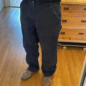Mörkblå Carhartt Byxor 34x32 - Flitigt använda byxor som trots det är i ett klart godkänt skick. Byxorna har en diskret ”fadeat” utseende vilket jag tycker gör dom snyggare, samt medelstora hål på vardera nedre baksida (se sista bilden). 