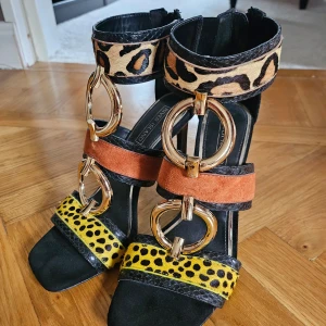 Färgglada klackskor med leopardmönster - Ett par eleganta klackskor från River Island med en unik design. Skorna har flera färger och mönster, inklusive leopardmönster på remmarna och guldfärgade ringdetaljer. De har en dragkedja på baksidan för enkel på- och avtagning.