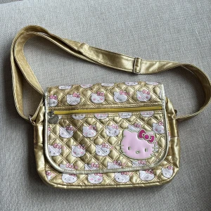 Väska Hello Kitty - Superfin väska i guld med Hello Kitty. Justerbar axelrem. Stort fack med dragkedja, litet fack med dragkedja på insidan. Fack på utsidan med dragkedja. Lite slitet på insidan men inget som syns utåt. Mått ca 32x23 cm
