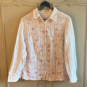 Vit Skjorta  - Vit skjorta med lila broderade mönster. Saknar storlek men skulle säga att den passar M-XL beroende på hur en vill att den ska sitta. Så fin och i bra skick men säljer för att den tyvärr inte kommer till användning. 