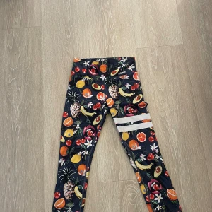 Färgglada leggings med fruktmönster - Säljer ett par supercoola leggings med ett färgglatt fruktmönster. De är svarta i grunden. Leggingsen har en hög midja och är tillverkade i ett stretchigt material som är perfekt för träning eller vardagsbruk. De har också två vita ränder på ena benet som en snygg detalj. 
