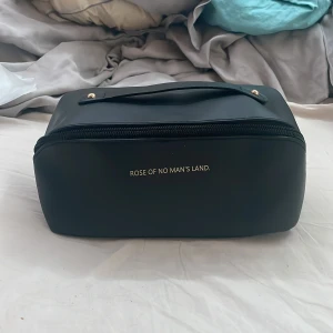 Svart necessär från Rose of No Man's Land - Säljer en snygg och praktisk svart necessär från Rose of No Man's Land. Den är perfekt för att förvara smink eller andra småsaker. Necessären har en dragkedja och ett handtag upptill för enkel transport. Insidan är rymlig med en vit foder och en extra ficka med dragkedja för att hålla saker organiserade. Perfekt för resor eller dagligt bruk! 
