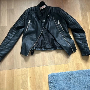 Svart skinnjacka med dragkedjor - Säljer en snygg svart skinnjacka i storlek S. Jackan har coola detaljer som dragkedjor på framsidan och quiltade partier på axlarna. Den är croppad och har spännen på baksidan för en justerbar passform. Perfekt för höst och vår!
