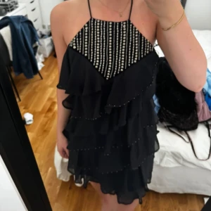Svart kortklänning med volanger - Säljer en superfin svart kortklänning med volanger och pärldetaljer. Klänningen har en öppen rygg med knytning. Perfekt för fest! Den är i nyskick och passar perfekt till sommarens alla fester.
