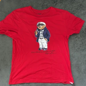 Röd t-shirt från Polo Ralph Lauren - Säljer en snygg röd t-shirt från Polo Ralph Lauren med en cool björn på framsidan. T-shirten är i bomull och har en normal passform. Perfekt för en avslappnad stil!