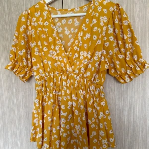 Gul blommig klänning - Säljer en superfin gul blus med vita blommor. Den har en v-ringad hals och korta puffärmar med resår. Blusen är i omlottstil och har en elastisk midja som ger en fin passform. Perfekt för sommarens alla fester och tillfällen! 🌼aldrig använd 