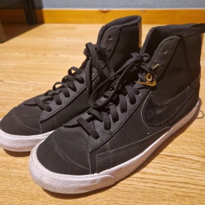 Svarta höga sneakers från Nike - Ett par svarta höga sneakers från Nike med vit sula och svarta snören. Skorna har en klassisk design med Nike-logotypen på sidan och en gulddetalj på hälen.