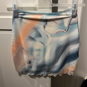 Mönstrad kjol från Gina Tricot - En kort kjol från Gina Tricot med ett abstrakt mönster i blå, orange och vita nyanser. Kjolen har en elastisk midja och är tillverkad i ett mjukt material.