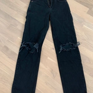 Svarta jeans med slitningar från Perfect Jeans - Säljer ett par svarta jeans från Perfect Jeans i storlek 34. De är högmidjade och har slitningar på knäna för en cool och avslappnad look. Jeansen är i bra skick och passar perfekt till både vardag och fest. De har en dragkedja och knapp framtill.
