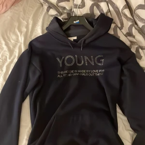 Svart hoodie från Gina Tricot - Säljer en snygg svart hoodie från Gina Tricot med texten 'YOUNG' i glittriga stenar på framsidan. Den är supermjuk och bekväm, perfekt för både vardag och chill. Hoodien har långa ärmar och en klassisk huva. Passar perfekt till jeans eller leggings!