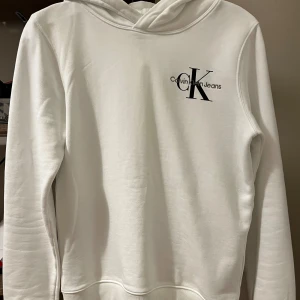 Vit hoodie från Calvin Klein Jeans - Säljer en snygg oanvänd vit hoodie från Calvin Klein Jeans. Den är i storlek 14 vilket är samma sak som storlek 164 och har en stor CK-logga på bröstet. Hoodien är gjord av mjuk bomull och har långa ärmar. Skriv om du är intresserad i den ❤️