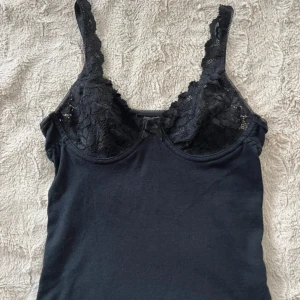Svart linne med spetsdetaljer - Säljer ett snyggt svart linne med spetsdetaljer på bysten. Perfekt för både vardag och fest! Linnet är ärmlöst och har en tight passform. Materialet känns mjukt och bekvämt mot huden.