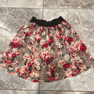 Blommig kjol med elastisk midja - En vacker kjol med ett blommigt mönster i rött, rosa och beige. Kjolen har en elastisk midja och är knälång.