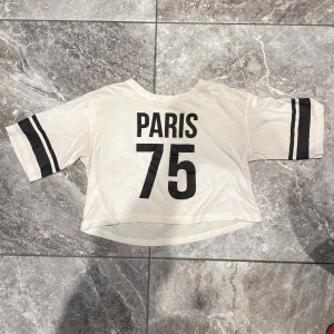 Vit topp med texten 'PARIS 75' - En vit topp med texten 'PARIS 75' i svart på framsidan. Toppen har korta ärmar med svarta ränder och en lös passform.