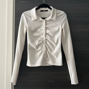 Vit skjort tröja  - Vit skjorta med långa ärmar och en knapprad framtill. Skjortan har en rynkad detalj på framsidan.