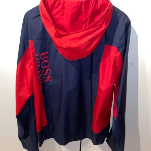 (Helt Ny) Blå och röd vindjacka från BOSS  - En stilren blå och röd vindjacka från BOSS med dragkedja framtill och huva. Jackan har röda detaljer på ärmarna och huvan samt BOSS-logotypen tryckt på bröstet och ryggen.