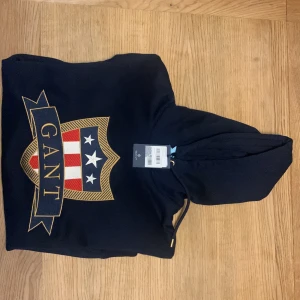 Oanvänd Mörkblå hoodie från Gant - Ny pris 899kr  En mörkblå hoodie från Gant med ett stort tryck av Gant-logotypen på framsidan. Hoodien har en justerbar huva och är tillverkad i ett mjukt material.