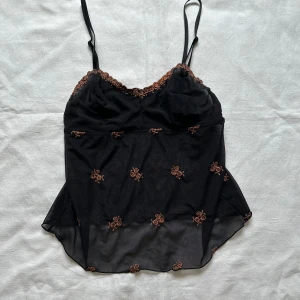 Svart linne med broderade blommor - Ett svart linne med tunna axelband och broderade blommor. Linnet har spetsdetaljer vid halsringningen och är tillverkat i ett genomskinligt material.