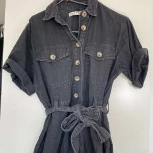 Svart jeansklänning från Denim & Co - En svart jeansbyxdress från Denim & Co med knappar framtill och två bröstfickor. Byxdresen har korta ärmar och ett knytskärp i midjan.