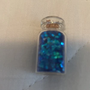 Litet glasflaska med blått glitter - En liten glasflaska fylld med blått glitter. Flaskan har en kork som förslutning.