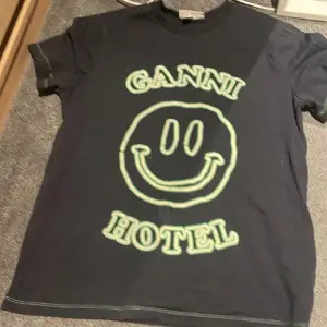 En svart t-shirt från Ganni med ett grönt smiley-ansikte och texten 'Ganni Hotel' på framsidan. Säljer den då den ej kommer till användning 