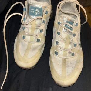 Nike Air Vapormax - Ett par vita Nike Air Vapormax sneakers med blå detaljer och snörning. Skorna har en genomskinlig sula och är i välanvänt skick. sulan finns inte i skorna heller