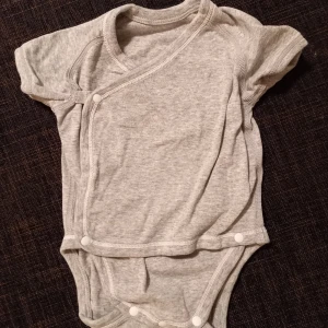 Grå t-shirtbody strl. 56/62 - Grå t-shirtbody, omlottbody, i storlek 56/62 (märkt 55-65 cm, 3-6 månader, 6 kg), från uniqlo. I fint skick, sparsamt använd. Inga hål eller fläckar. Unisex  Tvättad i parfymfritt tvättmedel och utan sköljmedel. 