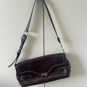 Brun väska  - Brun axelväska, med vita sömmar och silver detaljer. Rymlig med fack på insidan. Fint skick, dock tecken på användning.  36x19 cm utan axelband inräknat. 