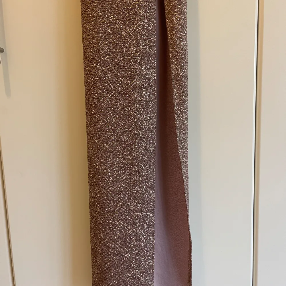 Lila och guldskimrande balklänning i stretchigt material. Hög slits, tunna axelband som korsas i ryggen. Några trådar har gått upp i ryggen men det är inget som syns tydligt (se sista närbilden). Normal i storleken, motsvarar storlek S-M då den är vä. Mekot.