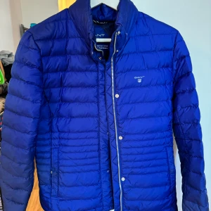 GANT jacka - Otroligt fin tunn jacka från GANT i dun. Blå färg men skimrar lite i ljuset. Perfekt som den är eller under en kappa. Med en tunn luva inbyggd i kragen. Aldrig använd! Hämtas i Täby Park eller mötes i Stockholm City! Kan även postas.