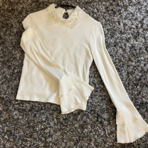 Blus med volang och pärlor  - Tajt blus med långa ärmar med volanger vid händerna. Kragen är full med pärlor vilket ger den perfekta looken. Superfin och dressig! 