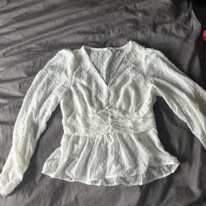 Vit blus med prickigt mönster - En elegant vit blus med ett diskret prickigt mönster. Blusen har långa ärmar och en v-ringad hals. Den är lätt och luftig, perfekt för både vardag och fest.