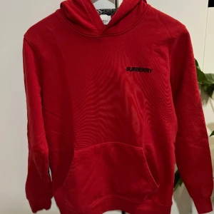 Burberry Hoodie - En Burberry Hoodie , i en härlig röd färg, väntar på att bli en del av din garderob! Denna klassiska och trendiga huvtröja är perfekt för att ge din outfit en touch av lyx och komfort.  Axelbred:42cm Längd: 68cm