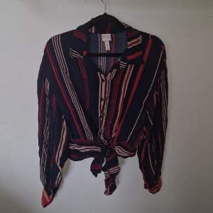 En randig skjorta (mörkblå, beige, röd) från H&M. Ballongärmar med breda muddar och knappar. Knytning nedtill med breda band. Aldrig använd.