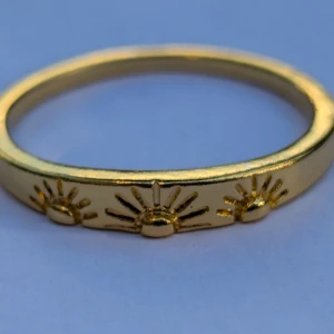 Guldfärgad ring med solmotiv - En elegant guldfärgad ring dekorerad med tre solmotiv. Ringen har en slät yta och en enkel, stilren design som passar för olika tillfällen.
