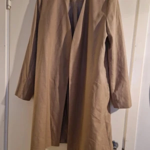 Beige kappa från Wera - En elegant beige kappa från Wera. Yttertyget består av 73% polyester, 19% viskos och 8% ull, medan fodret är gjort av 52% polyester och 48% viskos. Kappan har långa ärmar och en stilren design utan synliga knappar eller dragkedja. 