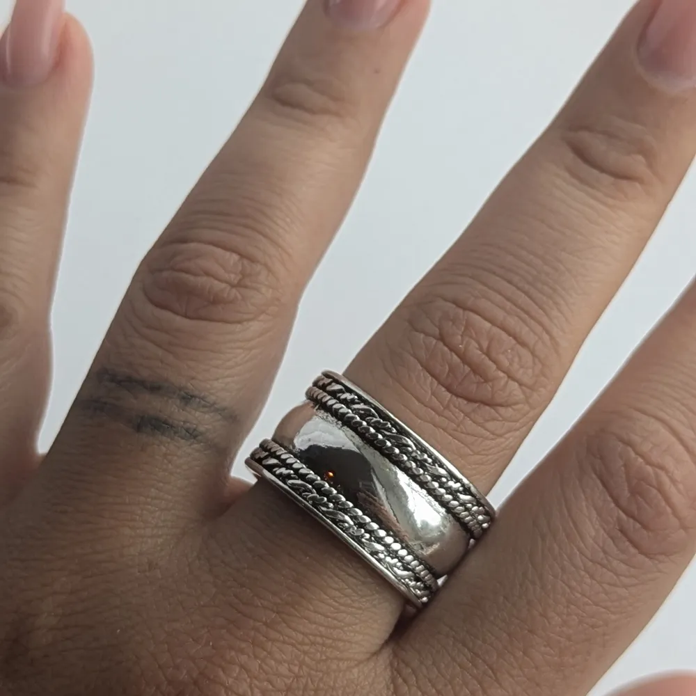 En bred silverring med en polerad yta och två flätade band som löper längs med ringens kanter. Ringen har en robust och elegant design. Tillverkad av koppar material, nytt. Accessoarer.