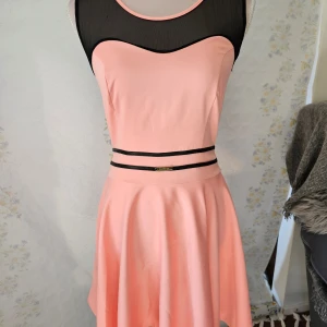 Rosa klänning från Maretto - En elegant rosa klänning från Maretto med svart mesh-detalj vid halsen och ryggen. Klänningen har en tight passform upptill och en utsvängd kjol. Den har även svarta banddetaljer i midjan.