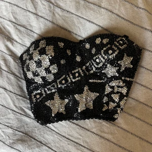 Svart tubtopp från Anna Tofana - En svart tubtopp från Anna Tofana dekorerad med silverfärgade paljetter i stjärnmönster. Toppen har justerbara axelband och en hak- och hyskstängning på baksidan. Har använts till blandannat cowboyfest och madonna outfit. Ena axelbandet e dock trasigt men går att använda utan! 