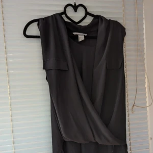 Svart ärmlös klänning - En elegant svart ärmlös klänning med omlott-design. Klänningen har en lös passform och är tillverkad i ett lätt och luftigt material. Den har en fickdetalj på bröstet och en asymmetrisk nederkant.