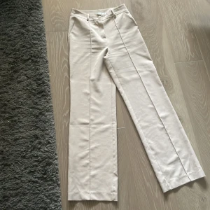 Beige kostymbyxor från Design by Si - Ett par beige kostymbyxor från Design by Si i storlek M. Byxorna har en hög midja och raka ben. De är tillverkade i ett mjukt och bekvämt material, perfekt för både formella och semi-formella tillfällen.